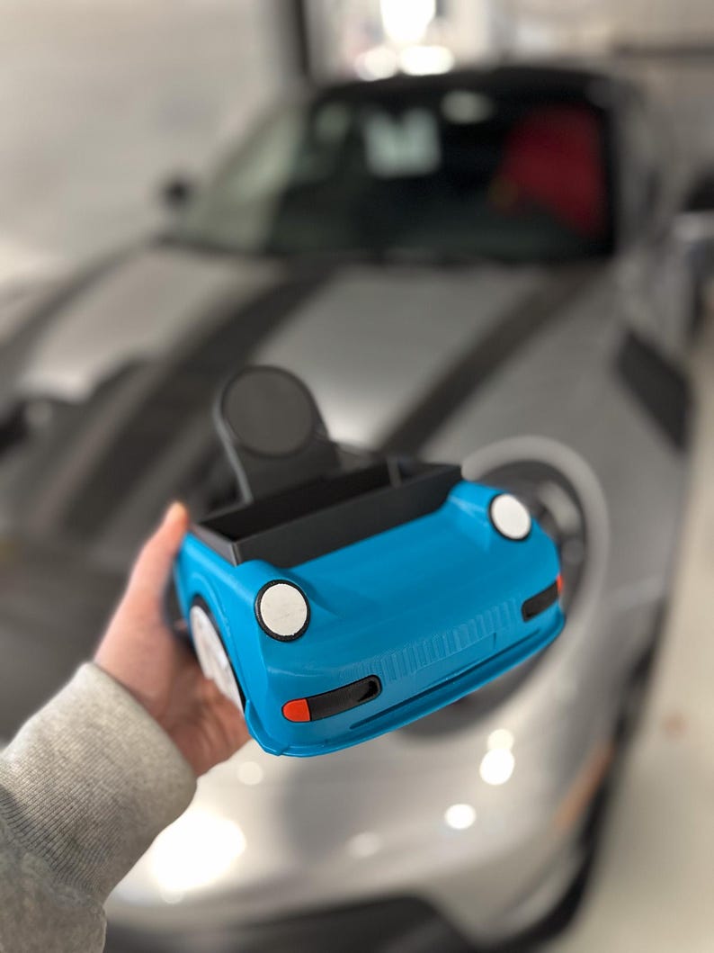 May include: A blue toy car with black accents and white headlights. The car is being held in a person's hand.