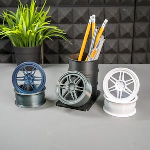 May include: Three small, 3D printed car wheels, one blue, one grey, and one white, are displayed on a table. A black cylinder with a grey rim holds a group of pencils and pens.