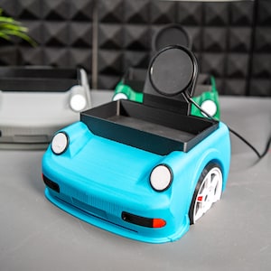 May include: A blue car-shaped phone stand with a black tray and a wireless charging pad. The stand has white headlights and a black rear bumper.