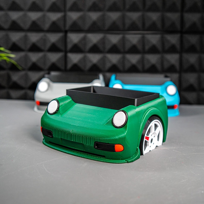 May include: Three car-shaped desk organizers, one green, one blue, and one gray. The green organizer is in focus and has a black interior.