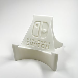 Peut inclure: Support en plastique blanc pour une console Nintendo Switch. Le support porte le logo Nintendo Switch et les mots "Nintendo Switch" imprimés dessus.