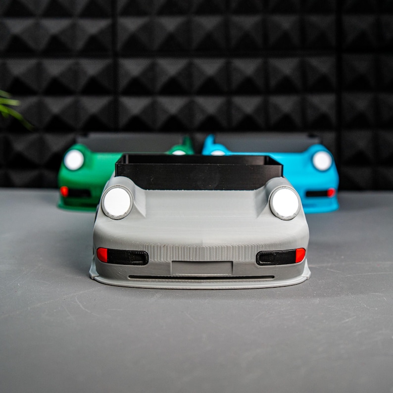 May include: Three gray, blue, and green car-shaped planters. The gray planter is in the foreground and has a black front grille and white headlights.
