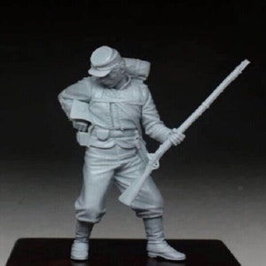 Op de afbeelding: Een grijs plastic model van een soldaat in uniform, met een geweer en een rugzak. De soldaat staat met zijn rechterbeen naar voren en zijn linkerarm gebogen achter zijn rug.
