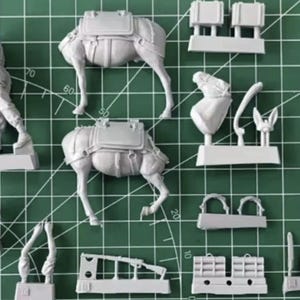 May include: A collection of unassembled plastic model pieces, including figures, camels with saddlebags, and various accessories. The parts are light gray and arranged on a green grid background.