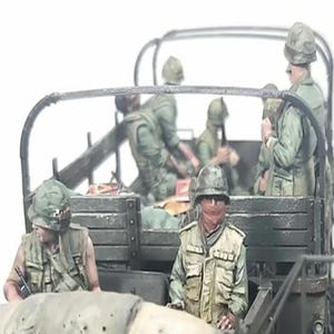 May include: A detailed diorama showing soldiers in a military vehicle. The figures wear green uniforms and helmets, with one wearing a red face covering. The vehicle has a wooden bed and a metal frame. The scene appears to be set during a conflict.