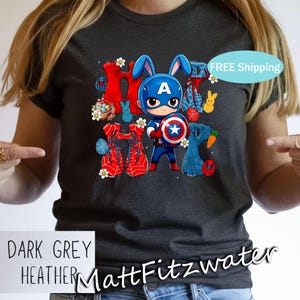 Pu&ograve; includere: T-shirt grigio scuro m&eacute;lange con un design pasquale a tema Captain America. La grafica include la parola "HOP" con elementi di supereroi e coniglietti. La maglietta ha anche il testo "MattFitzwater".