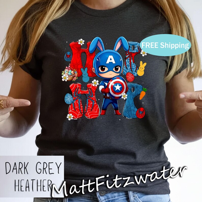 Pu&ograve; includere: T-shirt grigio scuro m&eacute;lange con un design pasquale a tema Captain America. La grafica include la parola "HOP" con elementi di supereroi e coniglietti. La maglietta ha anche il testo "MattFitzwater".
