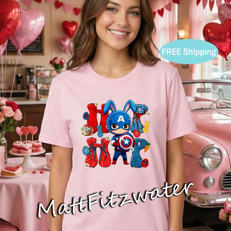 Pu&ograve; includere: T-shirt rosa chiaro con una grafica di un coniglietto vestito da Capitan America e la scritta "HOP" in rosso e blu. La maglietta ha girocollo e maniche corte. Lo sfondo comprende palloncini rosa e un'auto rosa.