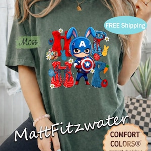 Pu&ograve; includere: T-shirt Comfort Colors verde scuro con una grafica di un coniglio supereroe e la scritta "Happy". Il design include uno scudo, orecchie da coniglio ed elementi a ragnatela. La maglietta ha un tocco morbido e vintage.