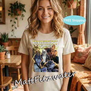 以下が含まれることがあります： ベージュ色のTシャツ。擬人化された猫の漫画の画像と、黄色と緑色の「PAWBERT Lynxley」の文字がプリントされています。アーティスト名「MattFitzwater」がTシャツの下部に印刷されています。