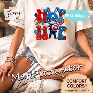 Pu&ograve; includere: T-shirt color avorio con una grafica di un coniglietto di Captain America e la parola "Hoppy" in rosso e blu. La maglietta ha un tocco morbido e vintage. Il testo "MattFitzwater" &egrave; visibile.