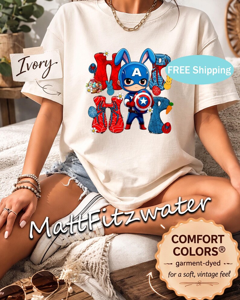 Pu&ograve; includere: T-shirt color avorio con una grafica di un coniglietto di Captain America e la parola "Hoppy" in rosso e blu. La maglietta ha un tocco morbido e vintage. Il testo "MattFitzwater" &egrave; visibile.