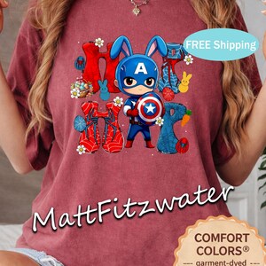 Pu&ograve; includere: Maglietta color cremisi con una grafica di un coniglio supereroe con elmetto di Capitan America e scudo. Il design include la parola "HOP" con elementi pasquali. La maglietta &egrave; tinta in capo per una sensazione morbida e vintage.