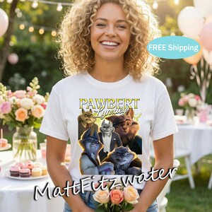 以下が含まれることがあります： 白いTシャツ。擬人化された猫のグラフィックと、黄色と緑の「PAWBERT Lynxley」の文字。Tシャツを着た人が花を持っています。背景には花とテーブルがあります。