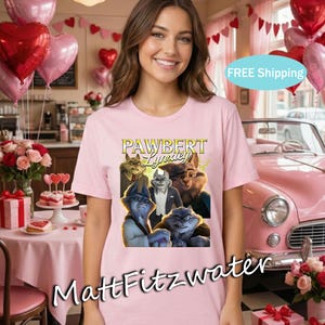 以下が含まれることがあります： 「PAWBERT Lyndsey」の文字と、スーツを着た擬人化された猫のグラフィックがプリントされた、明るいピンク色のTシャツ。カフェで、風船や花を背景に、そのTシャツを着ている人。