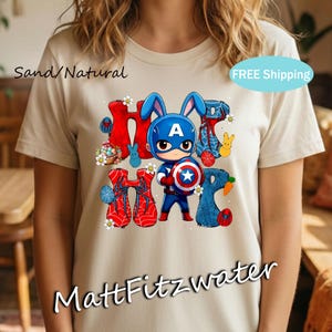 Pu&ograve; includere: T-shirt color sabbia con una grafica di un coniglietto supereroe che tiene uno scudo. Il design include la parola "Hoppy" in rosso e blu, con accenti a tema pasquale. La maglietta ha il testo "MattFitzwater" in basso.