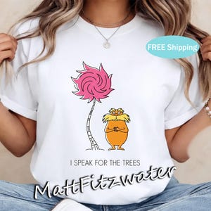 May include: White t-shirt featuring a cartoon illustration of a pink Truffula tree and the Lorax character. The text "I SPEAK FOR THE TREES" is printed below the image, with the artist's name at the bottom. A fun, whimsical design.