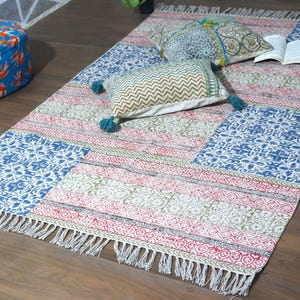 Può includere: Un tappeto rettangolare caratterizzato da un design patchwork con motivi floreali e geometrici stampati in blu, rosso e verde, rifinito con frange. Due cuscini decorativi con motivi coordinati e nappe poggiano sulla superficie.
