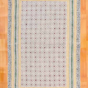 Puede incluir: Alfombra rectangular estampada con fondo crema y diseño floral repetido en rojo. La alfombra tiene un borde verde, amarillo y azul marino con flecos en los bordes. La alfombra está colocada sobre un suelo de madera.