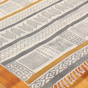 May include: A rectangular, patterned rug with a geometric design. The rug features stripes of white, gray, and gold. The design includes various shapes and patterns, with a fringed edge. The rug is on a wooden floor.