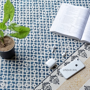 Puede incluir: Una alfombra con estampado blanco y azul con un borde amarillo y negro. Un libro, un teléfono y un par de auriculares inalámbricos están sobre la alfombra. El teléfono tiene el texto "@Nilesh Arts" en la pantalla.
