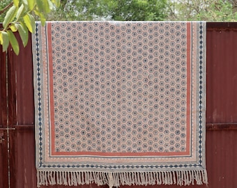 May include: A rectangular textile with a peach-coloured background and a repeating blue floral design. The border features a navy blue and cream diamond pattern, with a rust-coloured stripe. The bottom edge has a fringe.