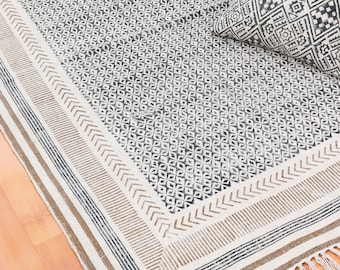 May include: A white and black patterned rug with a brown border and fringe. The rug has a geometric design and is made of cotton.