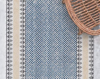 Könnte beinhalten: Ein rechteckiger Teppich mit einem zentralen blau-weißen Rautenmuster und Blumenmotiven. Der Teppich hat einen beigen Rand mit schwarzen Blumenakzenten und Fransen an den kurzen Seiten. Ein brauner Korbstuhl steht auf dem Teppich.