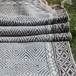 Pode incluir: Cobertor dobrado com padrão azul e branco com um design geométrico. O cobertor apresenta padrões de diamantes e ziguezague, com franjas na borda. A paleta de cores inclui tons de azul, branco e cinza.