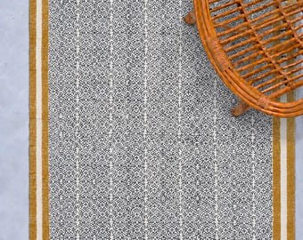 Puede incluir: Una alfombra rectangular con un panel central de patrón geométrico repetitivo en blanco y negro. La alfombra está enmarcada por un borde grueso de color amarillo mostaza y un borde interior blanco fino, con flecos color crema en los extremos.