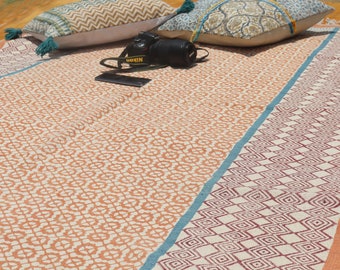 May include: A white and orange patterned rug with a teal border. The rug has a geometric design and is made of cotton. A camera is lying on the rug.