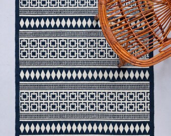 May include: A rectangular area rug featuring a dark navy blue and off-white geometric pattern with repeating square motifs and diamond shapes. The rug has a fringed edge on the top and bottom sides and a flat-woven texture throughout the surface.