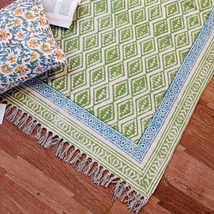 Könnte beinhalten: Ein grün-weiß gemusterter Teppich mit Rautenmuster und blauem Rand. Der Teppich hat an einer Kante Fransen. Ein Kissen mit Blumenmuster und ein Buch befinden sich in der oberen linken Ecke. Der Teppich liegt auf einem Holzboden.