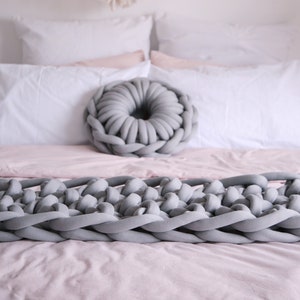 Könnte beinhalten: Eine graue gestrickte Wolldecke und ein graues, rundes Kissen aus Strick auf einem rosa Bett mit weißen Kissen.