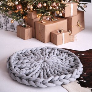 Könnte beinhalten: Ein großer grauer Strickhocker steht vor einem Weihnachtsbaum, der mit goldenen und rosafarbenen Kugeln geschmückt ist. Der Baum ist von verpackten Geschenken umgeben.