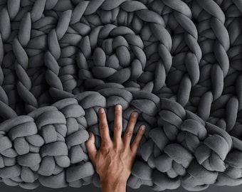 May include: A large, gray, chunky knit blanket made of thick yarn. The blanket is folded in half and a hand is reaching up to touch the top of the blanket.