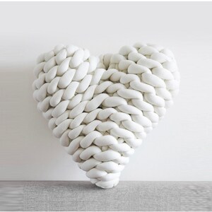 May include: A white heart-shaped pillow made of thick, braided fabric. The pillow is soft and fluffy, and it would be perfect for cuddling or decorating a bedroom.