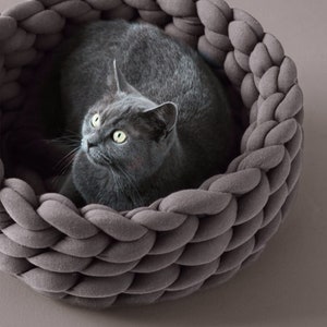 May include: A grey cat relaxing in a grey braided fabric pet bed. The bed is made of thick, woven fabric and has a round shape.