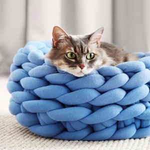 Op de afbeelding: Een grijze en witte kat met groene ogen rust in een blauw gevlochten dierenbed. Het bed is rond en gemaakt van dik, zacht materiaal. De kat kijkt recht in de camera.