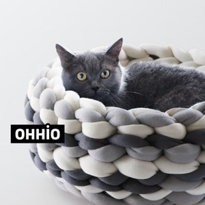 May include: A gray cat resting in a gray, white, and light gray braided pet bed. The bed is made of thick, woven fabric and has a round shape. The cat is looking directly at the camera with its yellow eyes. The text "OHHIO" is visible on the bed.