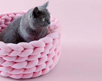 May include: A grey cat sitting in a pink woven pet bed.