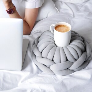 May include: A white ceramic mug filled with coffee or tea sits on a grey knitted pillow. The mug is on a white bed with a laptop and a person's arm in the background.