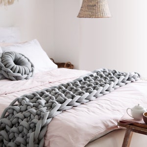 Puede incluir: Una manta de punto grueso de color gris sobre una cama rosa con almohadas blancas. La manta está hecha de hilo grueso y suave y tiene una sensación relajada y acogedora.
