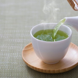 Puede incluir: Una tetera de cer&aacute;mica blanca vierte t&eacute; verde humeante en una taza blanca. La taza se apoya en un platillo peque&ntilde;o y redondo de madera. El t&eacute; es de un color verde vibrante, y sale vapor de la taza.