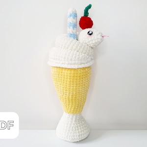 Puede incluir: Un juguete de peluche tejido a crochet con forma de batido, con una parte superior de crema batida blanca, una pajita a rayas amarilla y blanca y una cereza roja en la parte superior. El juguete está hecho de hilo suave y tiene un aspecto amigable y de dibujos animados.