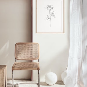 May include: A framed print featuring a minimalist line drawing of a rose. The artwork hangs above a wooden chair with a woven rattan back and seat. The chair has a chrome frame. A white sphere and vase are on the floor.