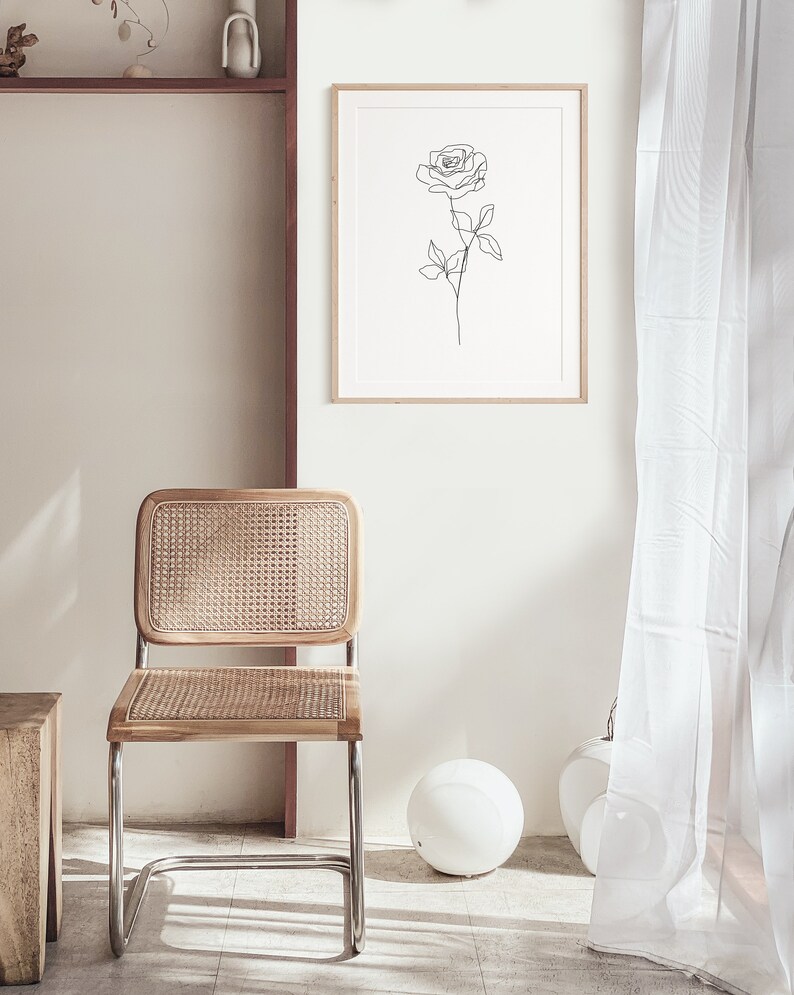 May include: A framed print featuring a minimalist line drawing of a rose. The artwork hangs above a wooden chair with a woven rattan back and seat. The chair has a chrome frame. A white sphere and vase are on the floor.