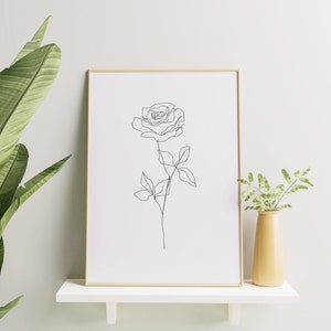 May include: A framed art print featuring a minimalist line drawing of a rose. The artwork is in a gold frame and displayed on a white shelf. A yellow vase with greenery is next to the frame, and a large green plant is on the left.