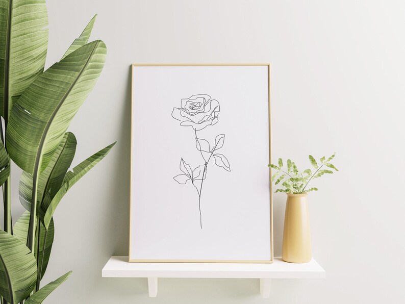 May include: A framed art print featuring a minimalist line drawing of a rose. The artwork is in a gold frame and displayed on a white shelf. A yellow vase with greenery is next to the frame, and a large green plant is on the left.