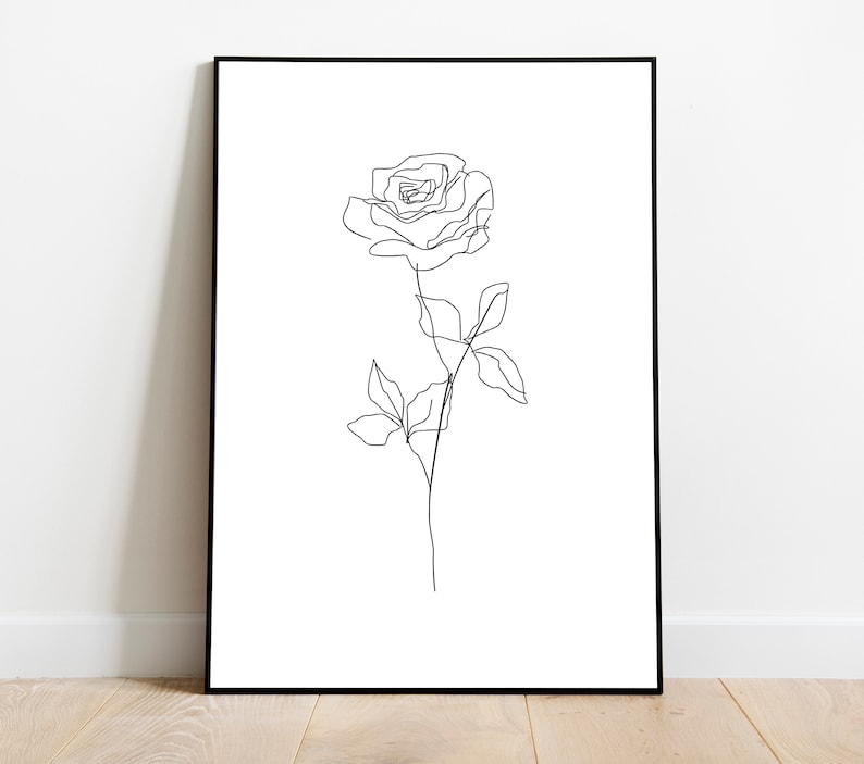 May include: Black framed art print featuring a minimalist line drawing of a rose. The artwork depicts a single rose with a stem and leaves, rendered in a continuous black line against a white background. The framed print is displayed on a white wall.